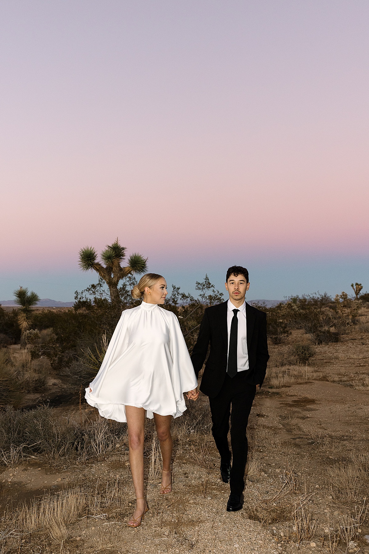 a bride in a white dress and groom in an all black suit at their Joshua Tree elopement during a purple and pink sunset